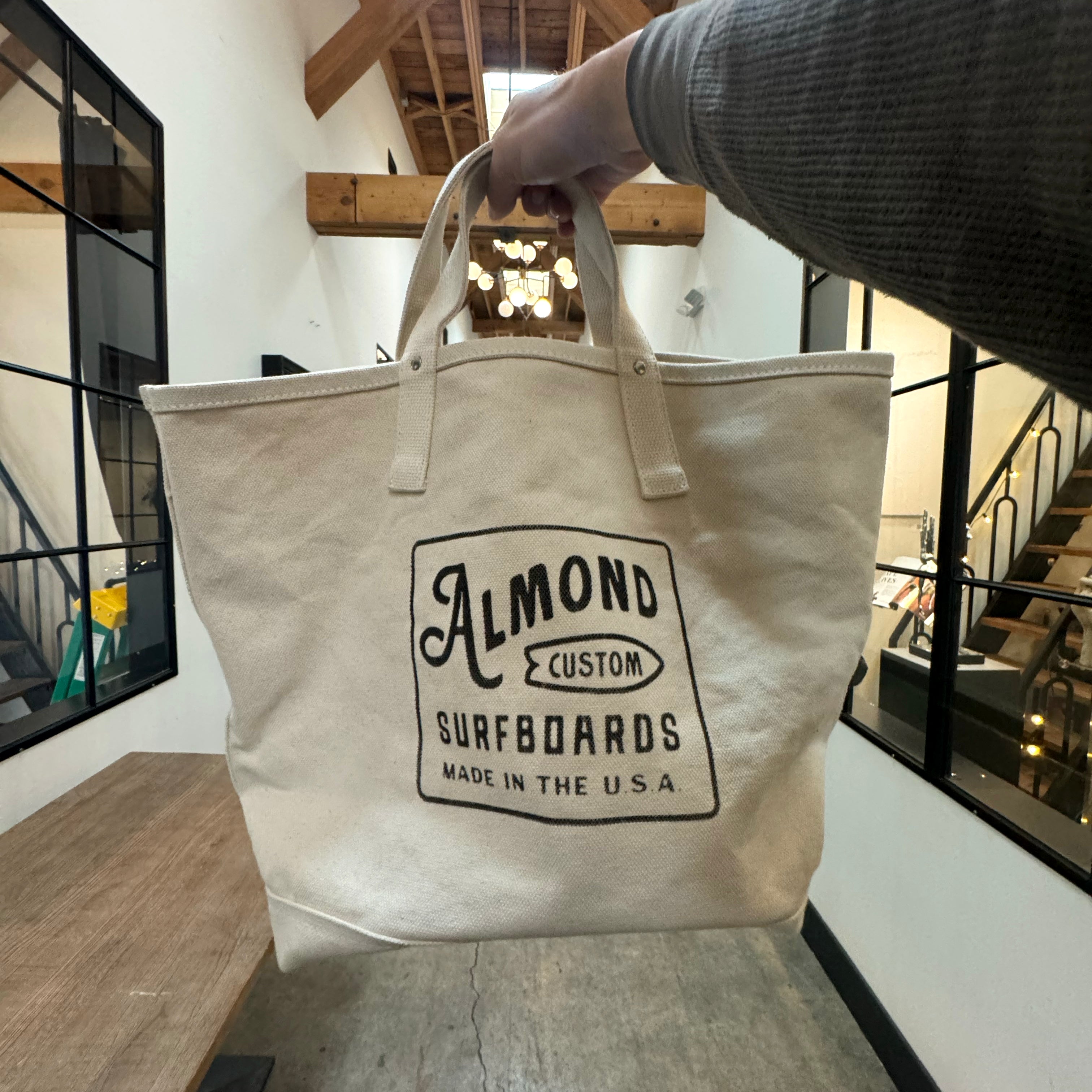 Beige tote bag with 'Almond Custom Surfboards' logo held by a person indoors.