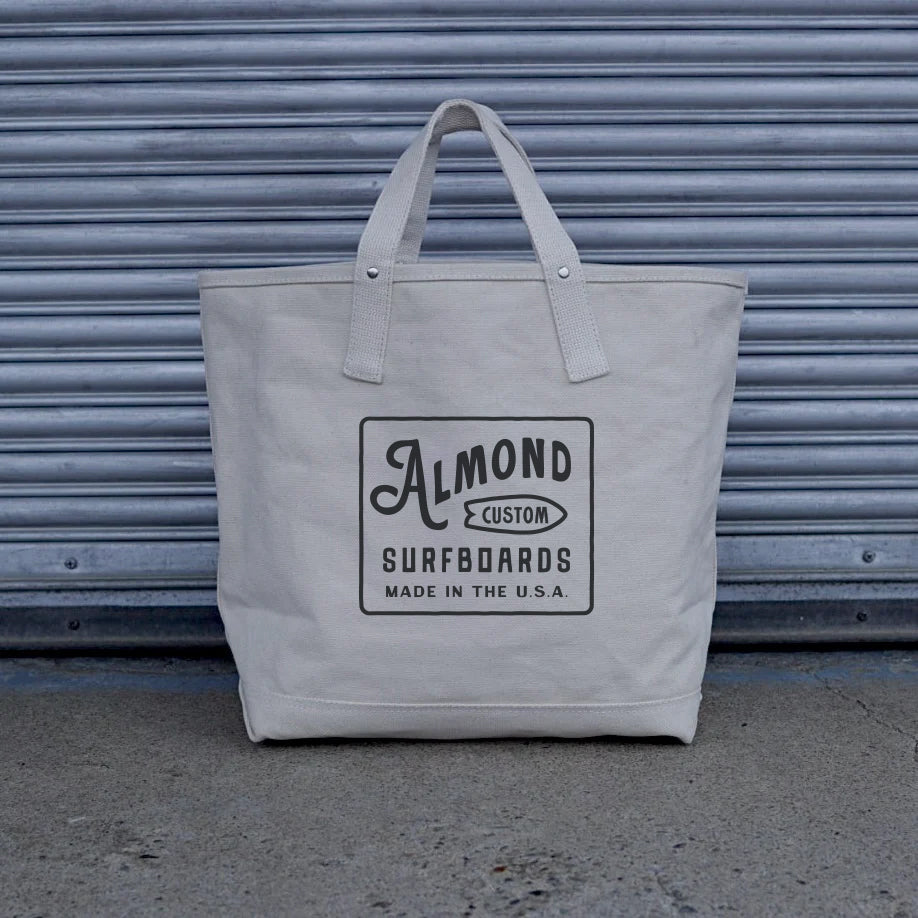 Gray tote bag with 'Almond Custom Surfboards' logo against a metallic shutter background