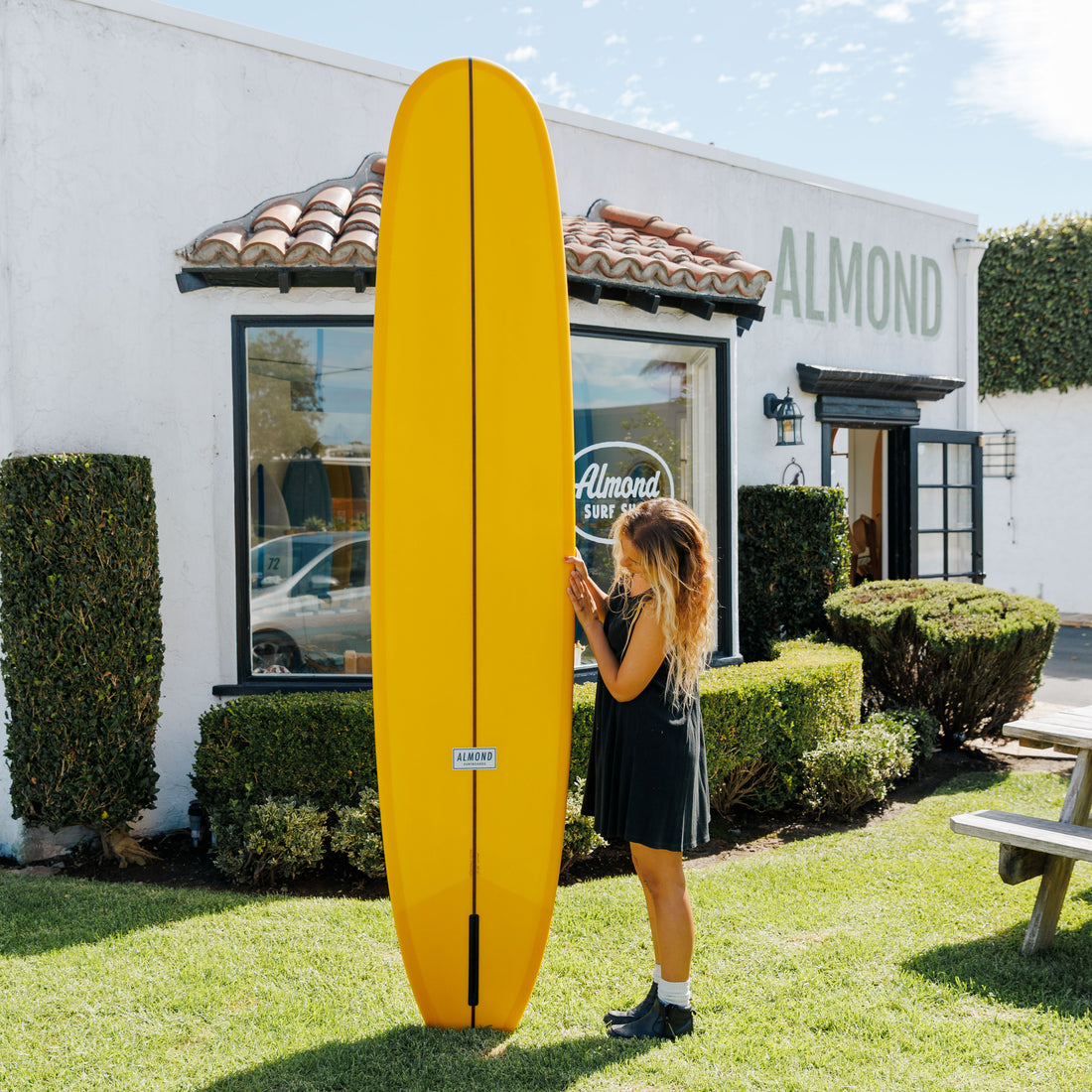 Person holding a yellow surfboard in front of a building with 'Almond' branding.