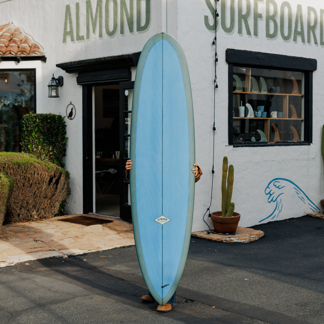 Person holding a blue surfboard in front of a building with 'Almond Surfboards' branding.