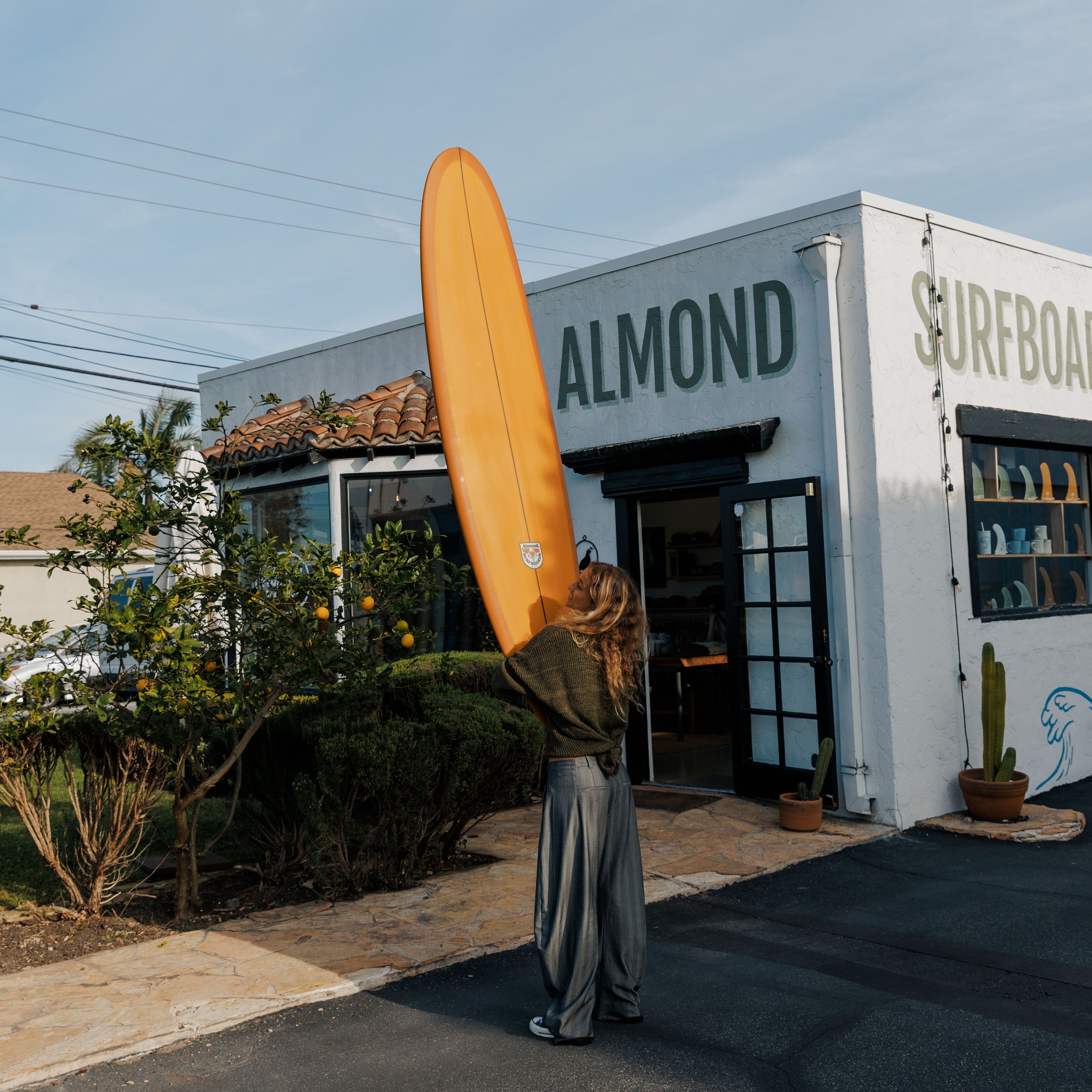 Person holding a surfboard outside a building with 'Almond Surfboards' branding.