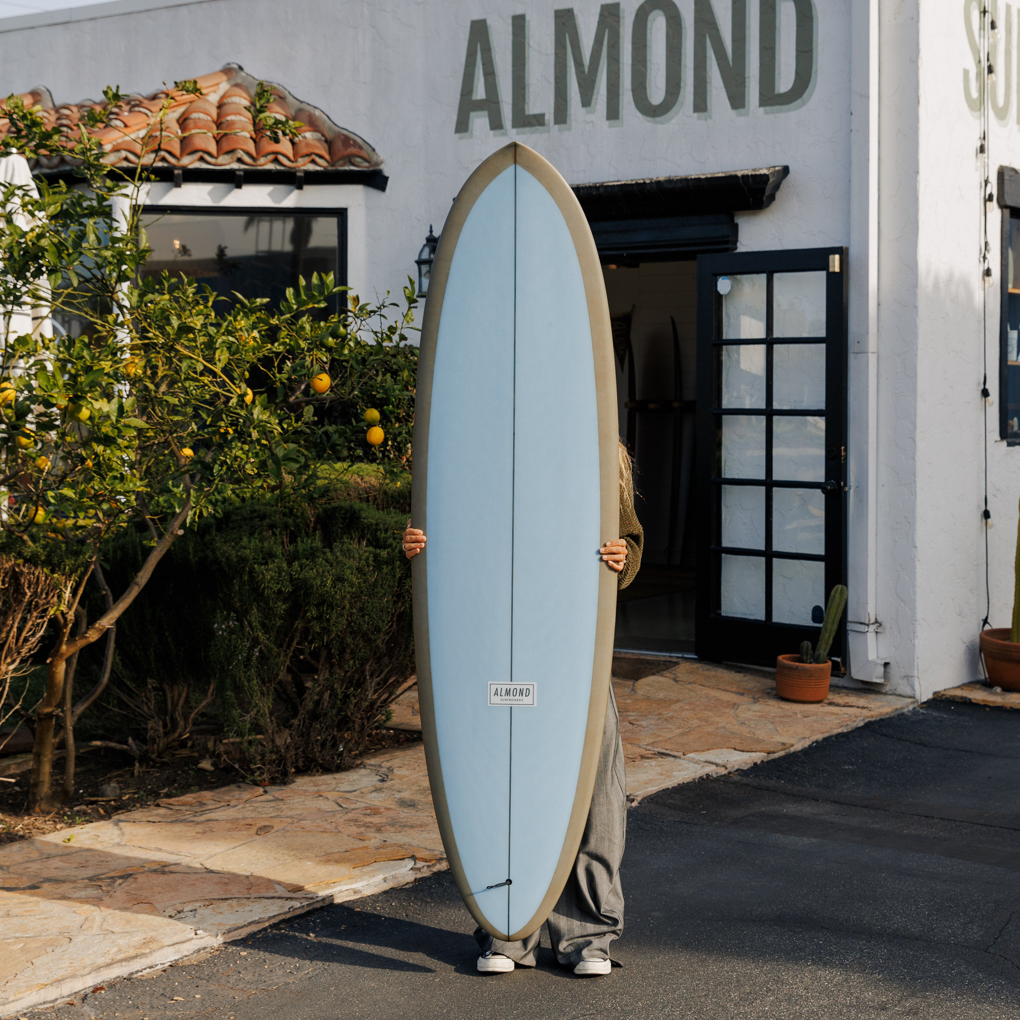 Person holding a surfboard in front of a building with 'ALMOND' branding.