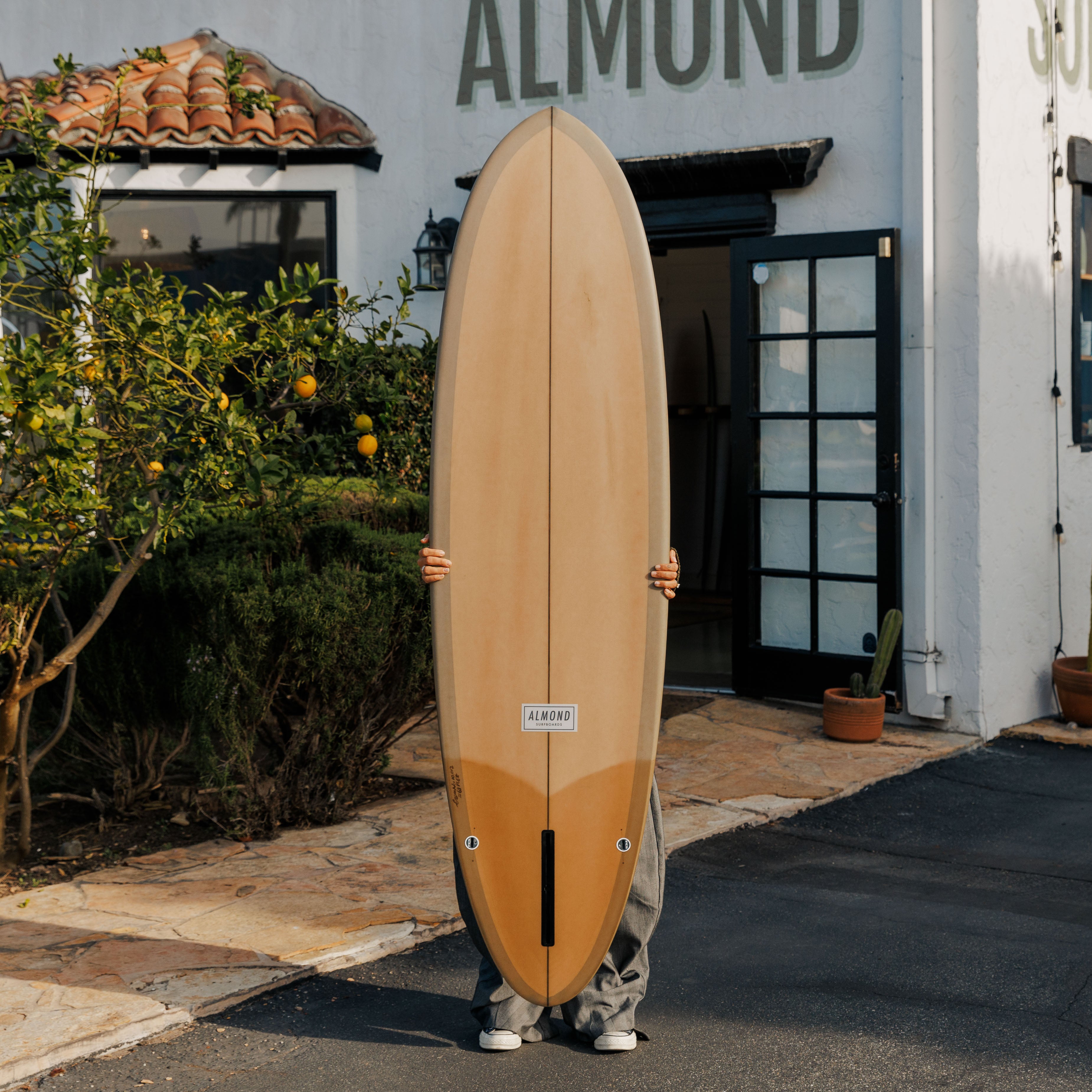 Person holding a surfboard in front of a building with 'ALMOND' branding.