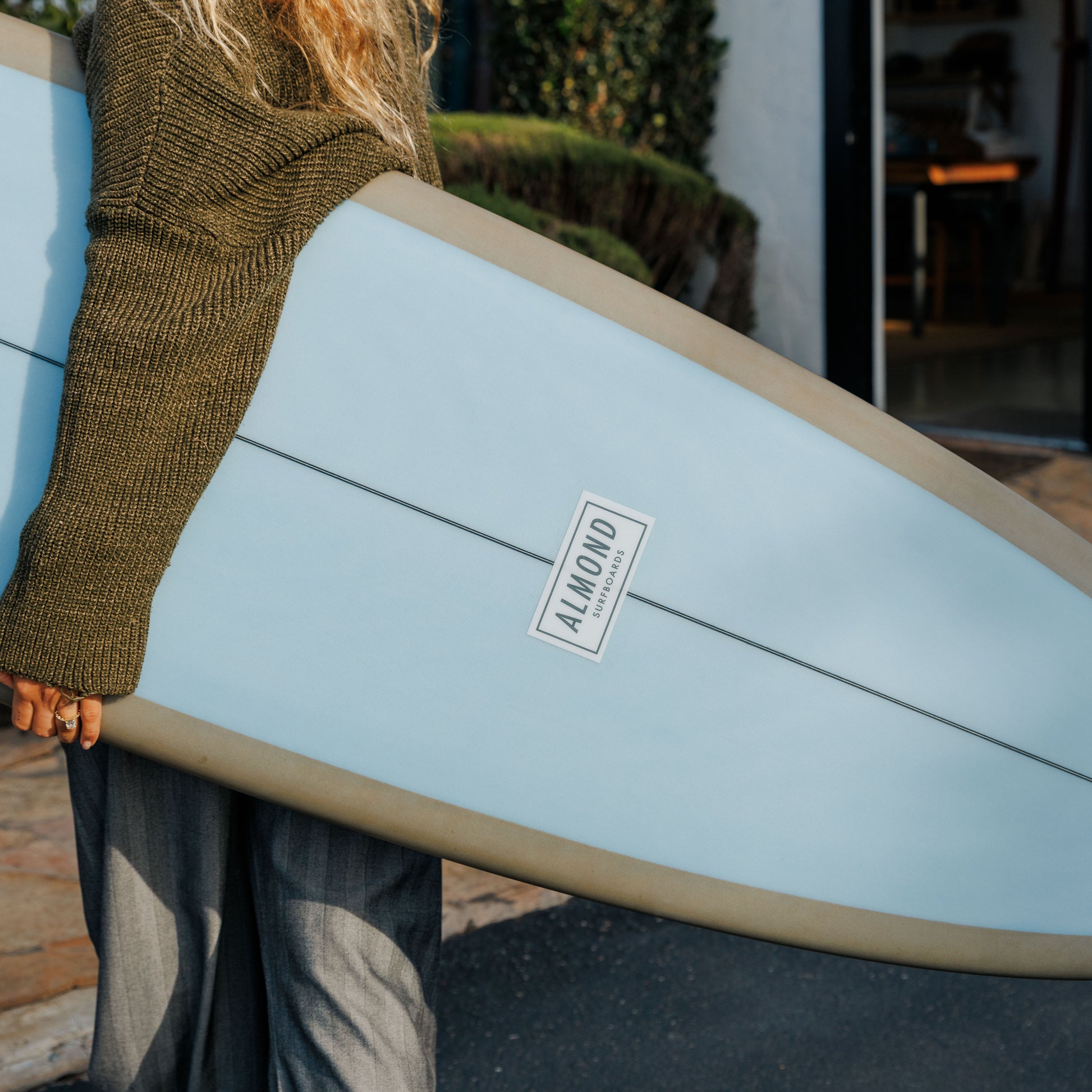 Person holding a light blue surfboard with 'Almond' branding outdoors.