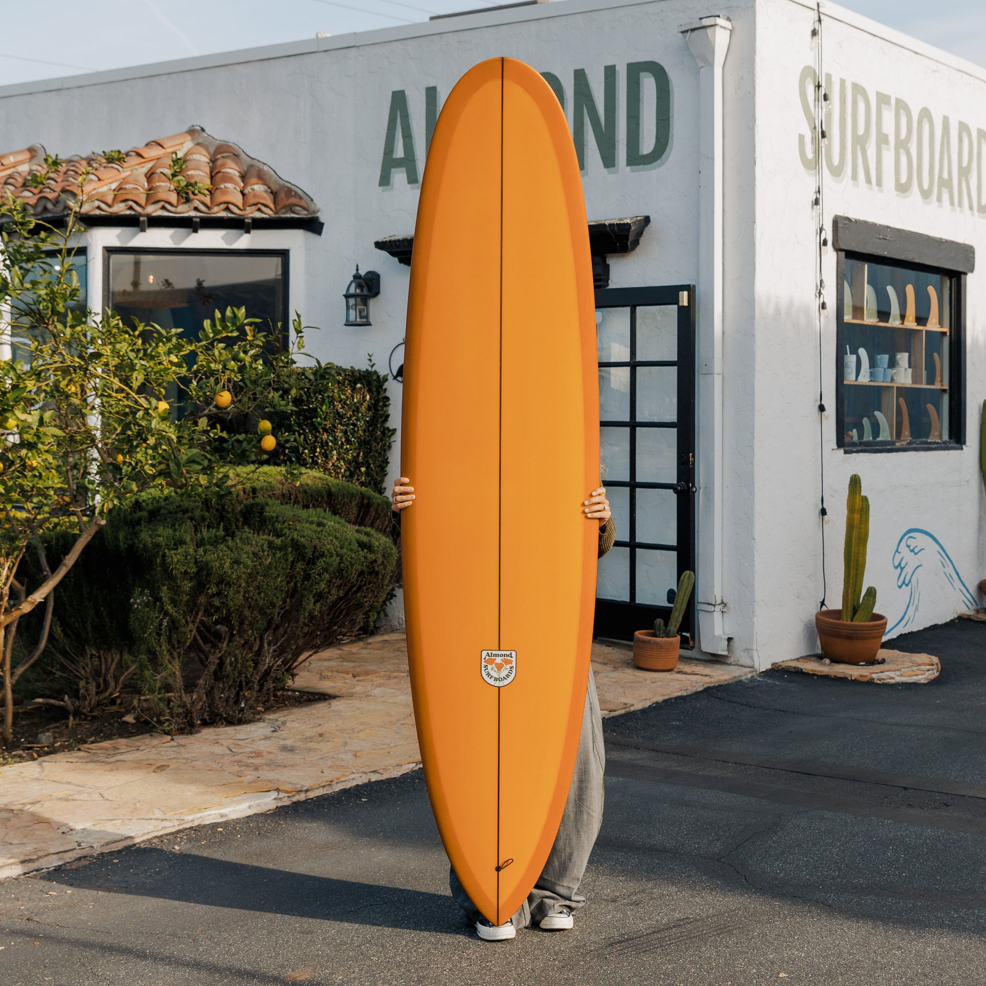 Person holding an orange surfboard in front of a building with 'Alond Surfboards' written on it.