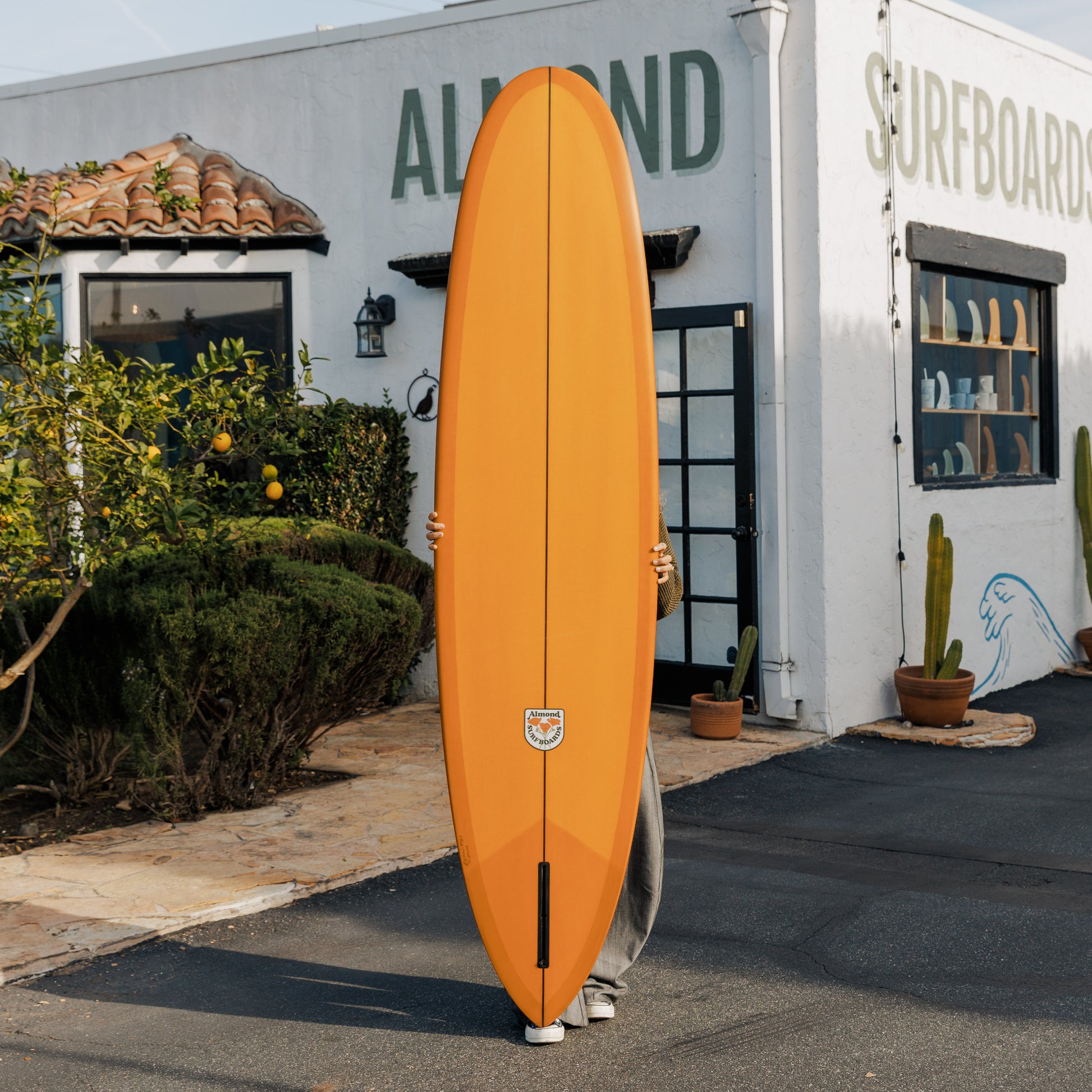 Person holding an orange surfboard in front of a building with 'Almond Surfboards' written on it.