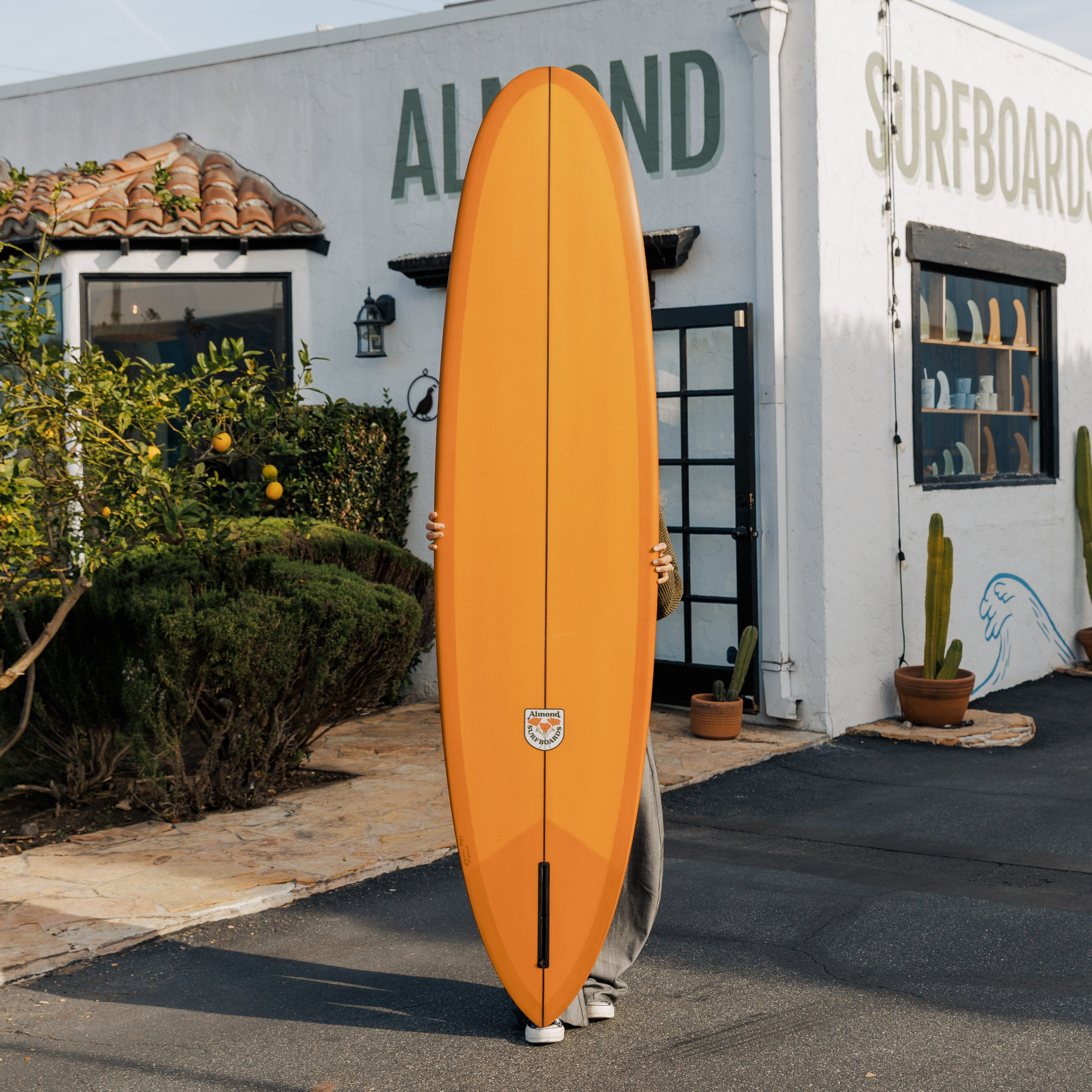 Person holding an orange surfboard in front of a building with 'Almond Surfboards' written on it.