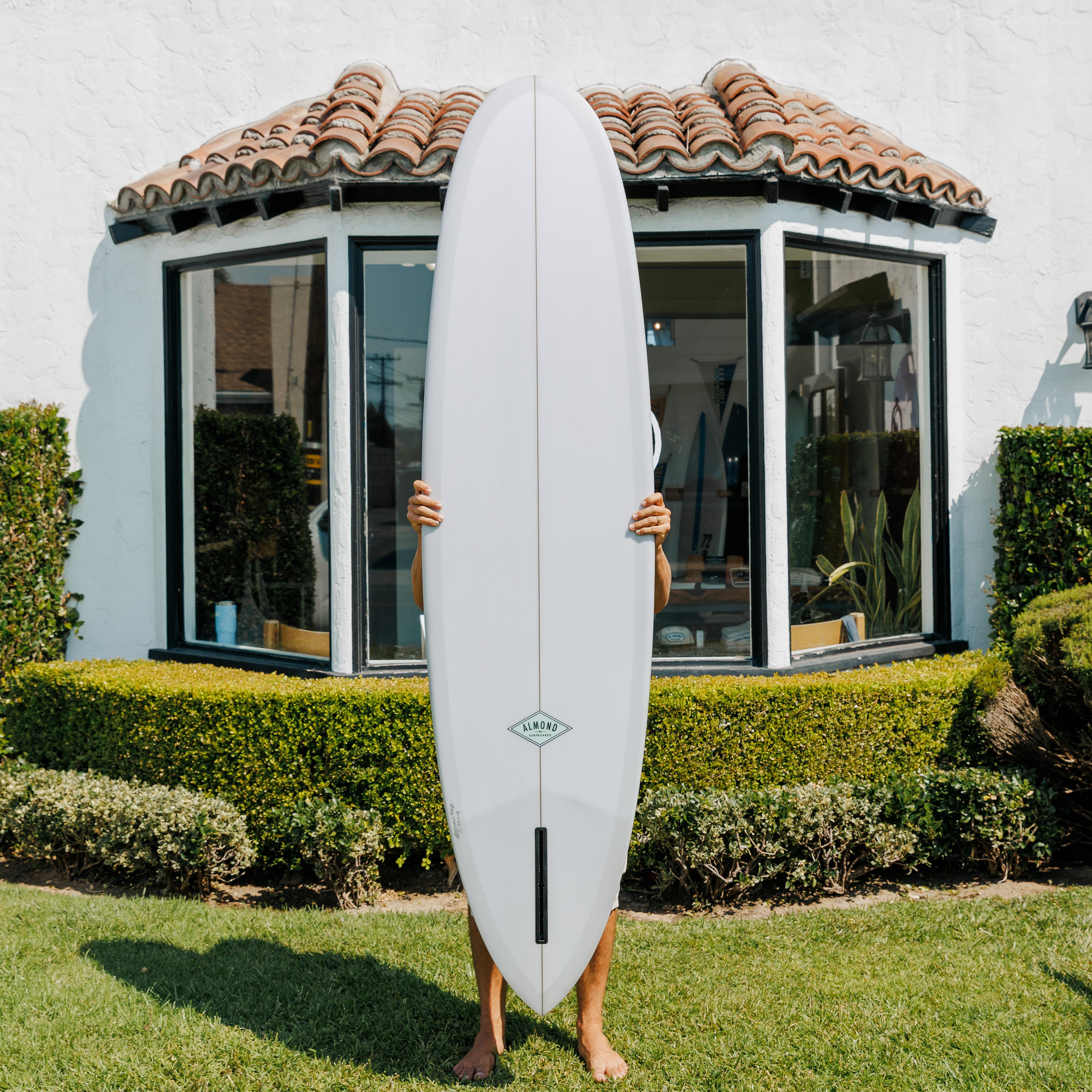 Person holding a surfboard in front of a house with a white exterior and red-tiled roof.